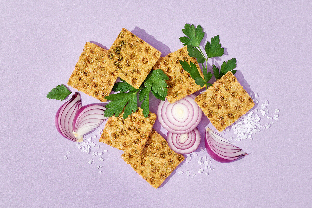 Knäckebrotsnack neben Petersilie, Zwiebalringen und Meersalz auf lila Untergrund, Foodfotografie