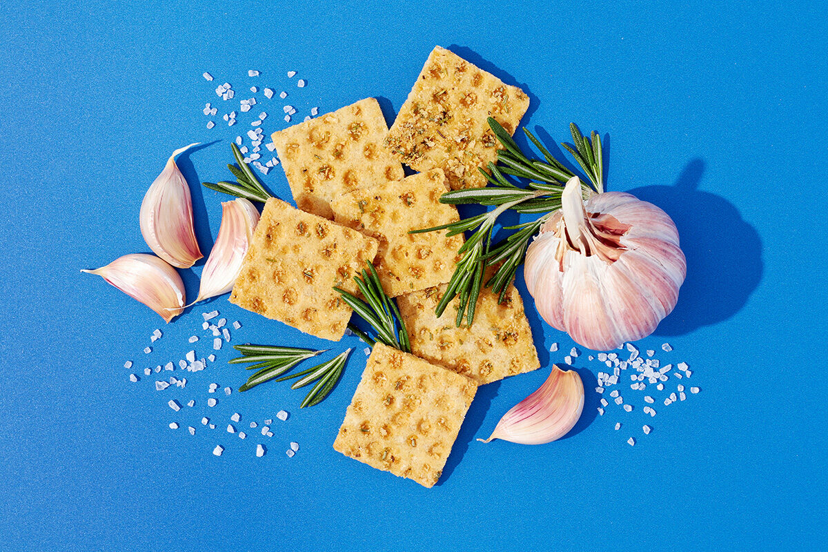 Knäckebrotsnack neben Knoblauch, Rosmarinzweigen und Meersalz auf blauem Untergrund, Foodfotografie