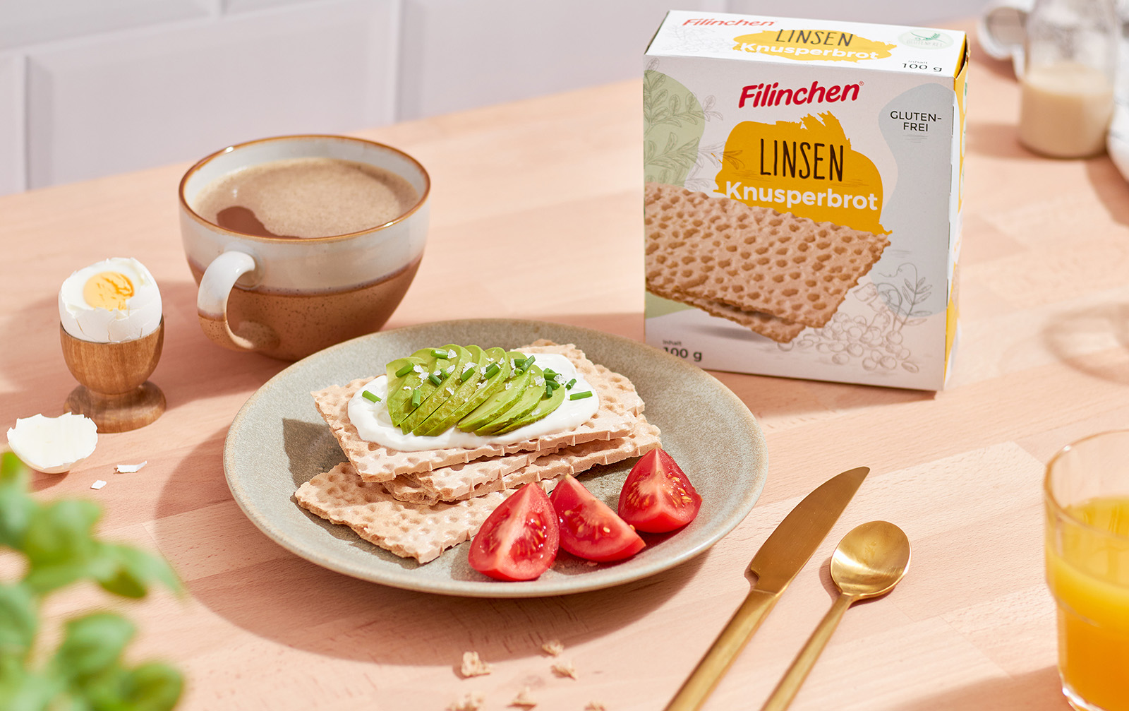 Teller mit Tomaten, Knäckebrotscheiben belegt mit Frischkäse und Avocado, daneben eine Tasse Kaffee, Besteck und die Verpackung des Knäckebrots, Foodfotografie