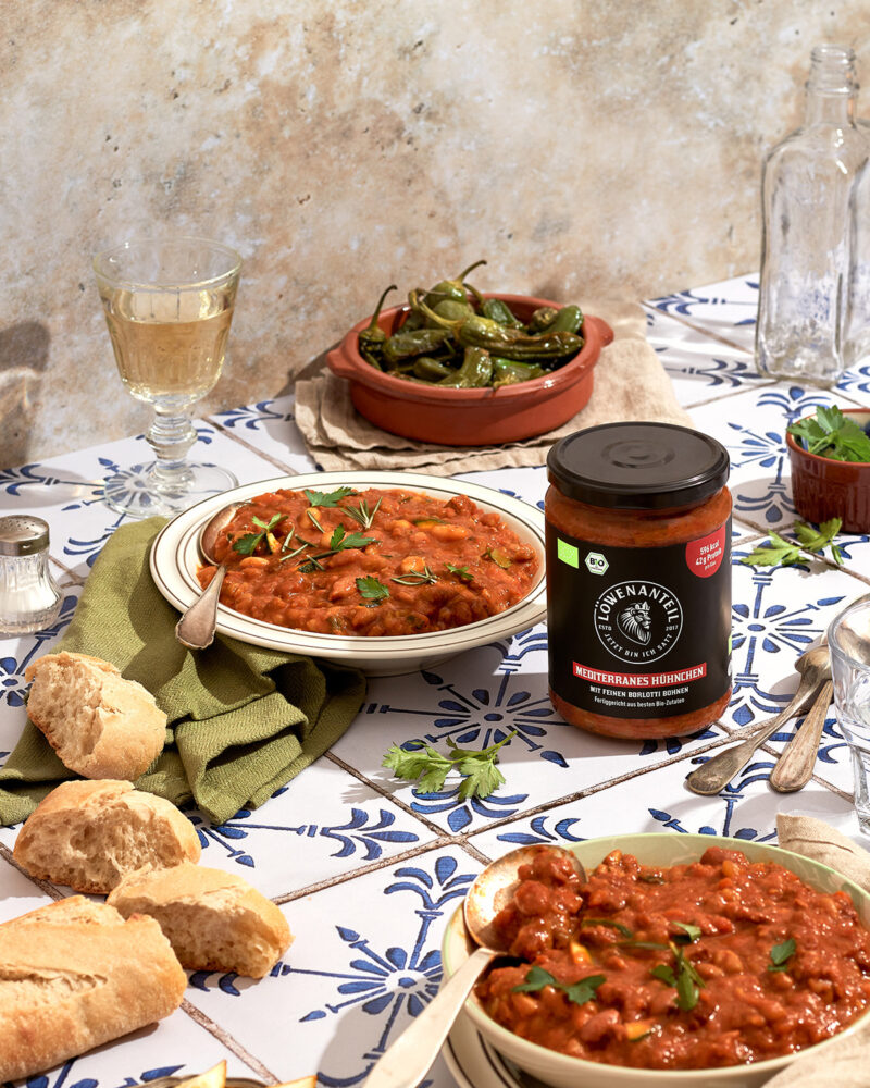 Glas Löwenanteil Sorte Mediterranes Hühnchen steht auf Kacheluntergund neben Teller mit Eintopf Foodfotografie